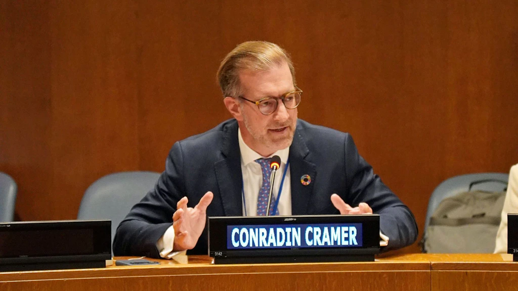 Conradin Cramer, wearing glasses and a suit, is speaking and gesturing at a conference table. There is a nameplate in front of him. An interpreting booth can be seen in the background.