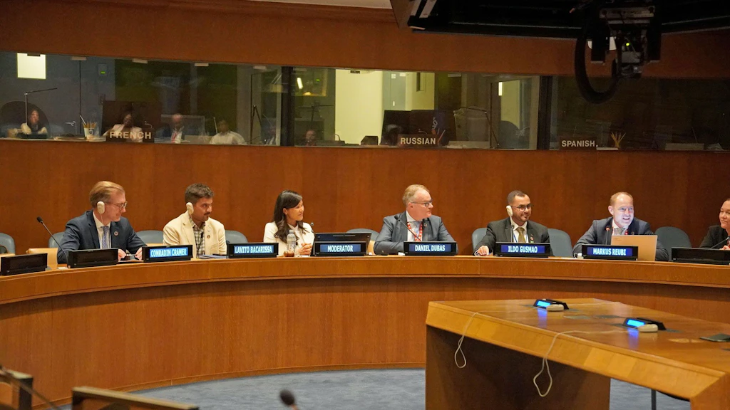 Seven people seated at a semi-circular table. Name tags in front of them. Interpreters’ booths in the background.