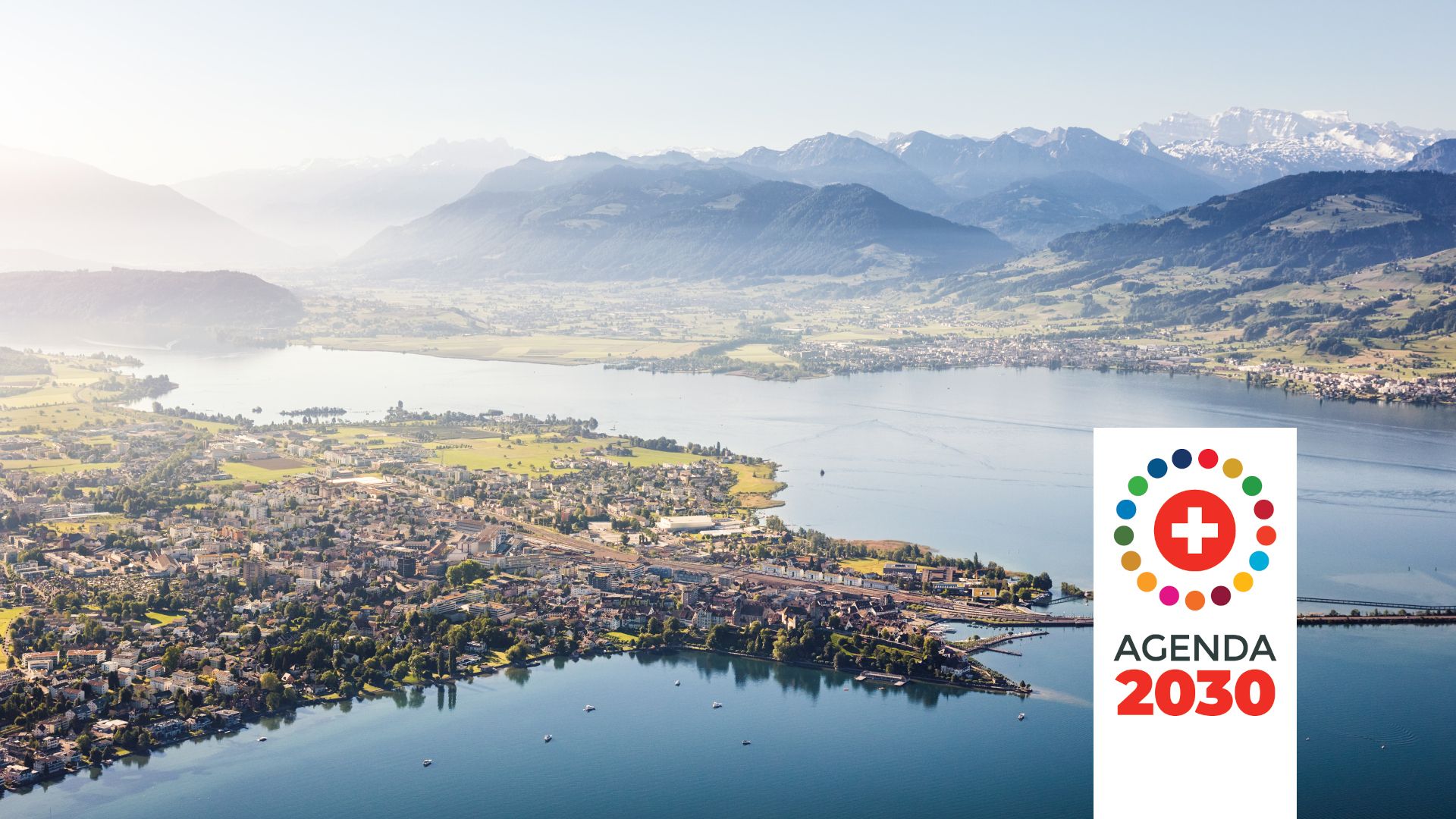 Un paysage typiquement suisse avec une prairie, un village, un lac et des montagnes, surmonté d'un logo avec la croix suisse entourée de points et de l'inscription « Agenda 2030 ».